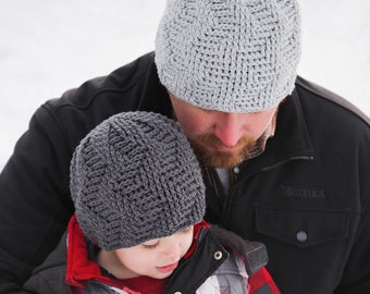 May include: A father and son wearing matching gray knitted hats. The father is wearing a black jacket with the word "Marmot" on it. The son is wearing a red and black plaid shirt under a red jacket.