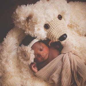 May include: A newborn baby wrapped in a beige blanket, wearing a floral headband, and resting on a large, fluffy, cream-colored teddy bear. The teddy bear has a crocheted face with brown eyes and a black nose.