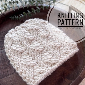 May include: Cream-colored knitted hat with a cable knit pattern. The hat is displayed on a wooden tray. Text on the image reads "Knitting Pattern" and "Jennifer Dougherty".