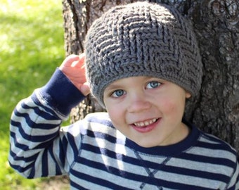 May include: A young child wearing a gray knitted hat with a braided pattern and a striped blue and gray sweater. The child is standing next to a tree.