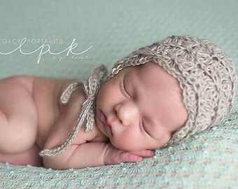May include: A newborn baby wearing a crocheted hat with a light brown color, sleeping on a light blue textured blanket. The baby is curled up with their arms and legs tucked in.