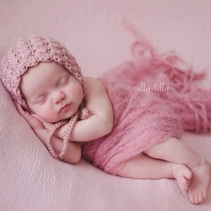 May include: A sleeping baby girl wearing a pink crocheted hat and a pink fuzzy wrap. The baby is lying on a pink background.