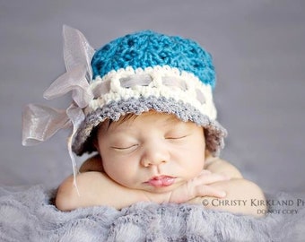 May include: A sleeping baby wearing a blue, white, and gray crocheted hat with a white satin bow. The baby is lying on a gray fuzzy blanket.