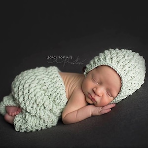 Puede incluir: Un bebé recién nacido con un gorro y pantalones de punto de color verde claro a juego, durmiendo sobre una superficie gris oscuro.