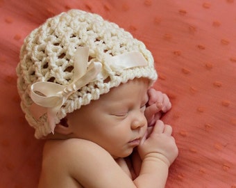 May include: A cream-colored crocheted baby hat featuring an open-weave pattern and a smooth, matching satin ribbon tied into a bow on the side. The hat is worn by a sleeping infant. The text Christy Kirkland Photography Do Not Copy is visible.