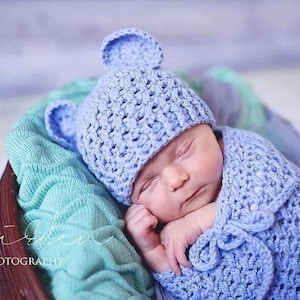 May include: A baby asleep, wearing a light blue crocheted hat with bear ears and a matching wrap. The baby is in a wooden basket on a turquoise blanket. The hat and wrap are made of a textured yarn, perfect for a newborn.
