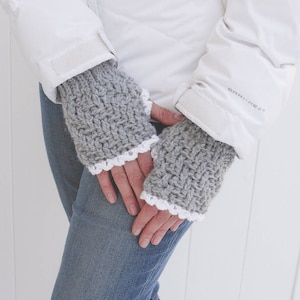 May include: Gray crocheted fingerless gloves with a white lace trim. The gloves are worn by a person wearing a white jacket and blue jeans.