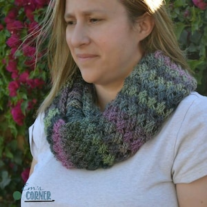Puede incluir: Primer plano de una mujer con una bufanda infinita de ganchillo en tonos verdes, morados y rosas. La bufanda está hecha con un punto de textura.