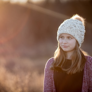 K&ouml;nnte beinhalten: Eine junge Person tr&auml;gt eine wei&szlig;e Strickm&uuml;tze mit einem flauschigen Bommel, einen lila Cardigan und ein schwarzes Shirt. Die Person schaut zur Seite und hat lange braune Haare.