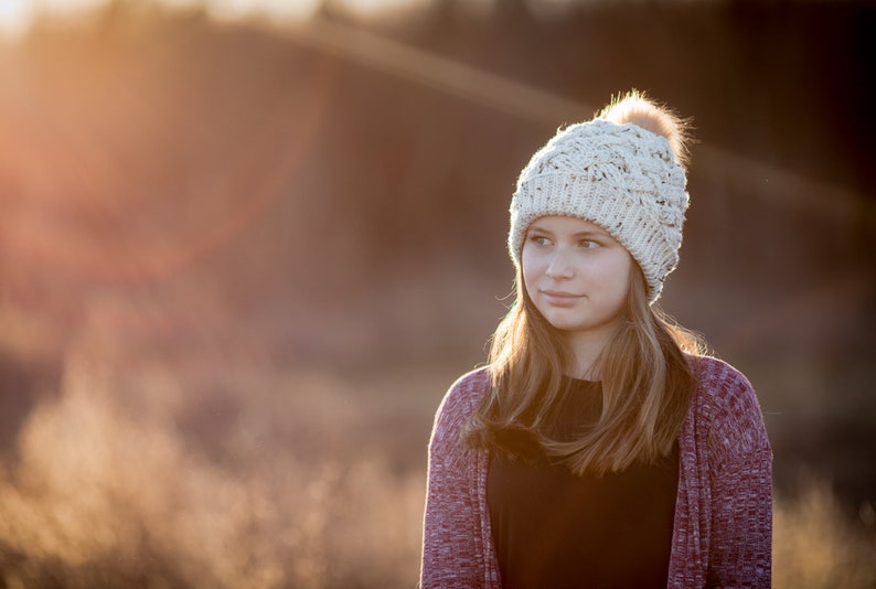 K&ouml;nnte beinhalten: Eine junge Person tr&auml;gt eine wei&szlig;e Strickm&uuml;tze mit einem flauschigen Bommel, einen lila Cardigan und ein schwarzes Shirt. Die Person schaut zur Seite und hat lange braune Haare.