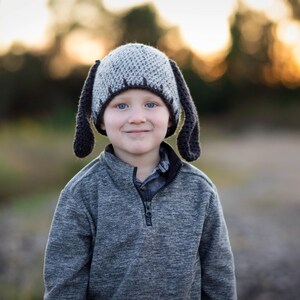 May include: A child wearing a gray and black crocheted dog hat with long floppy ears. The child is also wearing a gray zip-up sweater. The background is blurred with warm tones.