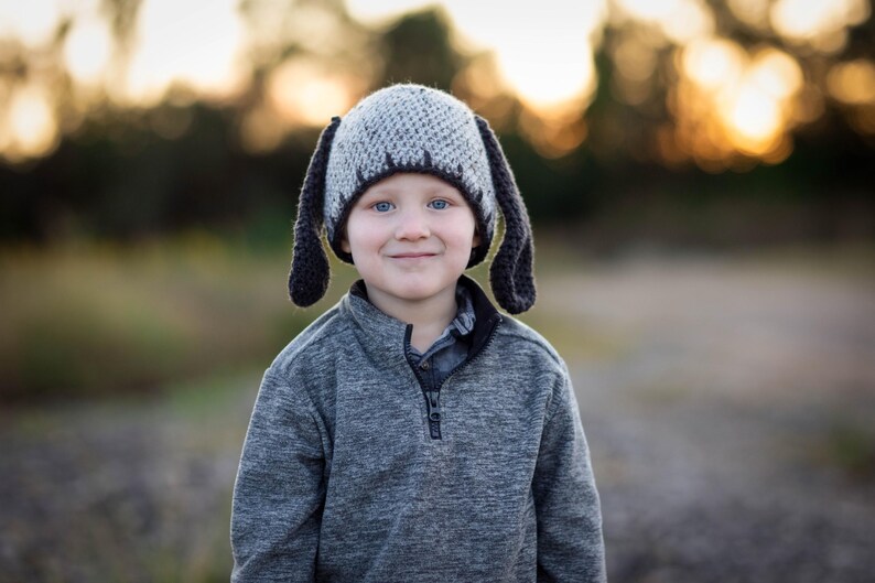 May include: A child wearing a gray and black crocheted dog hat with long floppy ears. The child is also wearing a gray zip-up sweater. The background is blurred with warm tones.