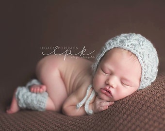 May include: A newborn baby wearing a light blue crocheted hat and leg warmers, sleeping on a brown textured surface. The baby is curled up with their hands under their chin.