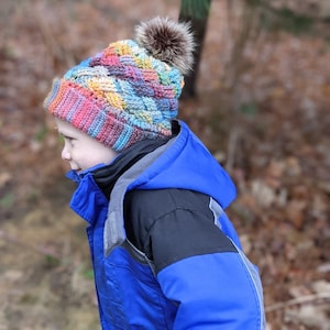 K&ouml;nnte beinhalten: Ein Kind tr&auml;gt eine bunte geh&auml;kelte M&uuml;tze mit einem braunen Bommel und eine blaue und schwarze Winterjacke.