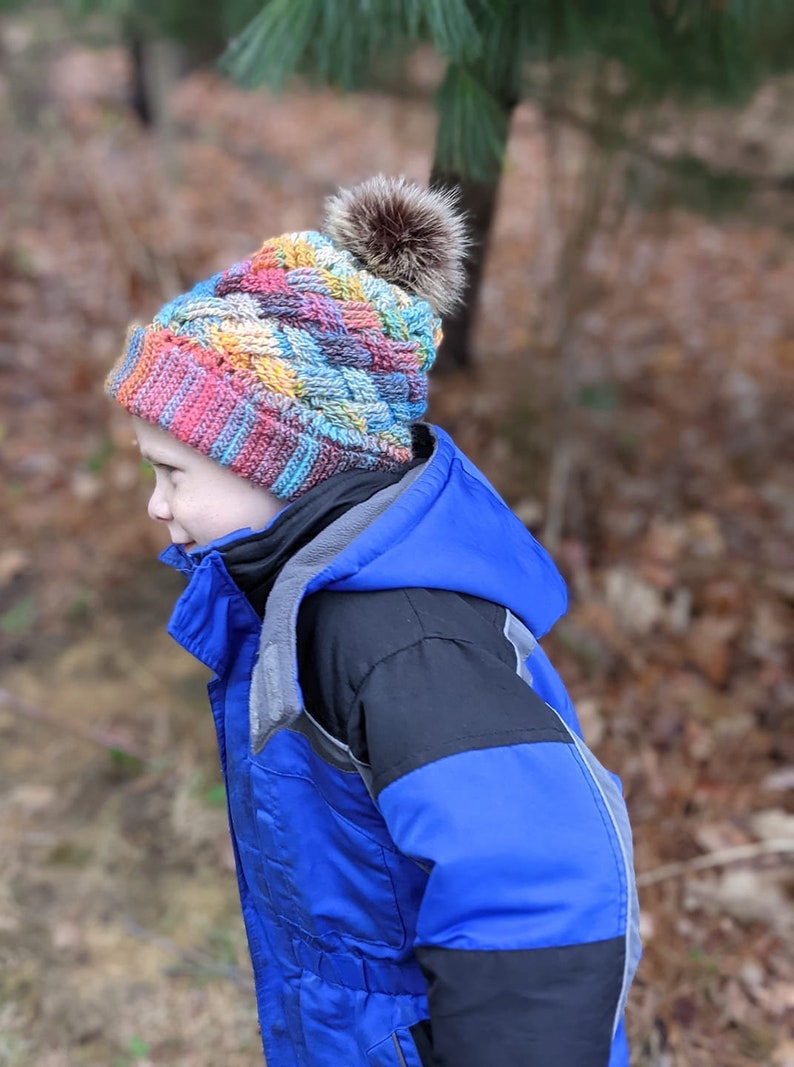 K&ouml;nnte beinhalten: Ein Kind tr&auml;gt eine bunte geh&auml;kelte M&uuml;tze mit einem braunen Bommel und eine blaue und schwarze Winterjacke.