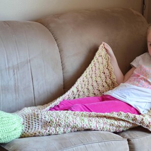 Op de afbeelding: Een gehaakte zeemeerminstaartdeken in lichtgroen en een cr&egrave;me- en roze kleurenschema. De deken is gedrapeerd over een persoon die een roze broek en een wit t-shirt draagt, rustend op een beige bank.