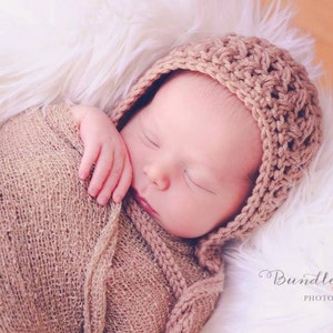May include: A newborn baby is sleeping soundly in a brown crocheted hat and a brown knitted wrap. The baby is lying on a white fluffy surface.