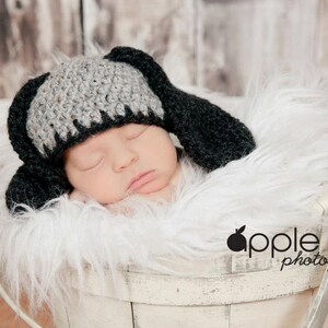 May include: A newborn baby wearing a handmade crochet hat with long black ears, resting in a white basket filled with fluffy white faux fur. The hat is gray and black. The background is a blurred wooden surface.