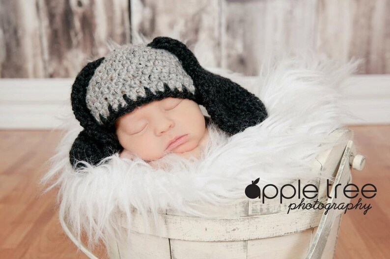 May include: A newborn baby wearing a handmade crochet hat with long black ears, resting in a white basket filled with fluffy white faux fur. The hat is gray and black. The background is a blurred wooden surface.