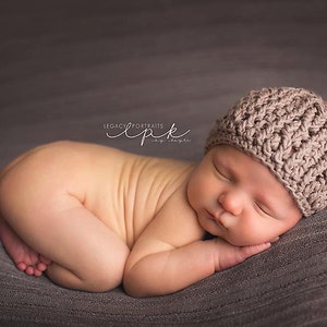 Puede incluir: Un bebé recién nacido con un gorro de punto marrón está durmiendo sobre una tela gris texturizada. El bebé está acurrucado con los brazos y las piernas metidos.
