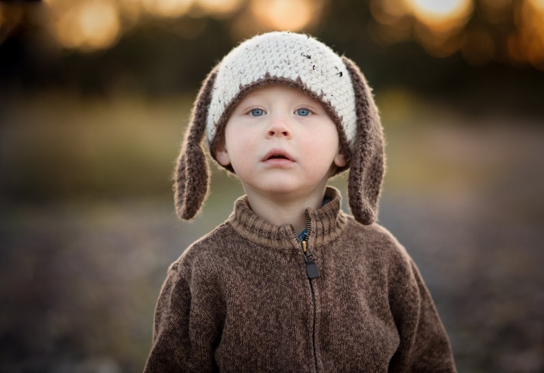 May include: A child wearing a handmade crochet hat with long, floppy ears. The hat is white with brown flecks and brown ear details. The child is also wearing a brown zip-up sweater.