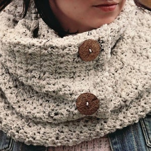 May include: A white crocheted infinity scarf with a speckled pattern and two wooden buttons.
