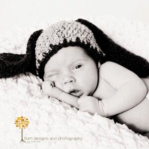 May include: A newborn baby wearing a black and gray crocheted hat with long floppy ears, lying on a soft, white, textured blanket. The baby is winking and resting their head on their hand. The image is in black and white.