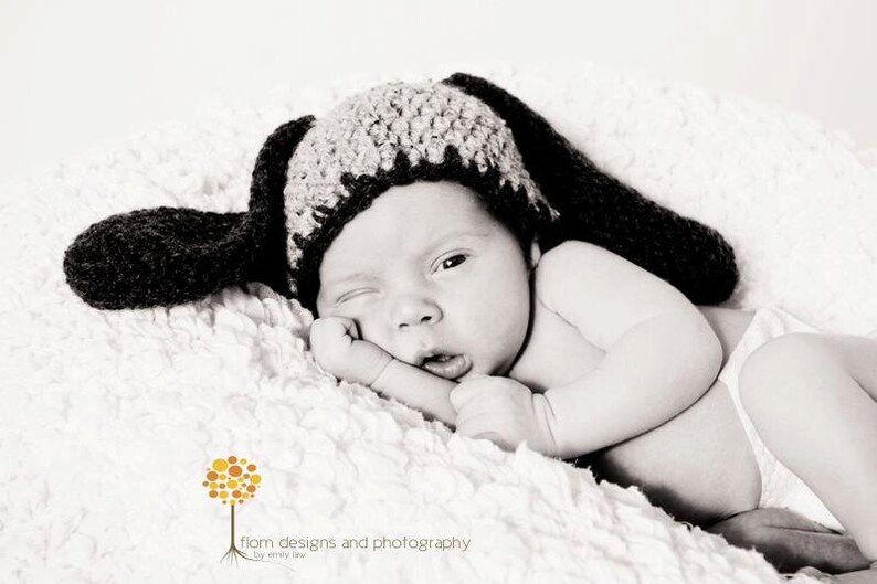 May include: A newborn baby wearing a black and gray crocheted hat with long floppy ears, lying on a soft, white, textured blanket. The baby is winking and resting their head on their hand. The image is in black and white.