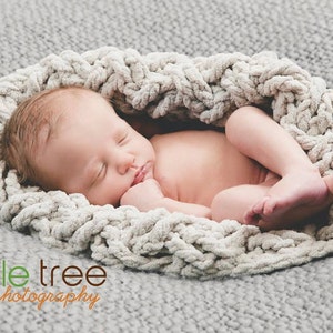 Könnte beinhalten: Ein neugeborenes Baby schläft friedlich in einer weichen, hellgrauen, grob gestrickten Decke. Das Baby liegt mittig im Bild, die Decke bildet ein gemütliches, kreisförmiges Nest. Der Hintergrund ist ein passender grauer Stoff.