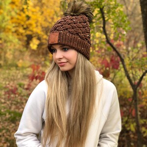 K&ouml;nnte beinhalten: Eine braune Strickm&uuml;tze mit einem Kunstfellbommel und einem braunen Lederpatch mit der Umrandung des Bundesstaates Minnesota. Die M&uuml;tze wird von einer Person mit langen blonden Haaren und einem wei&szlig;en Hoodie getragen.