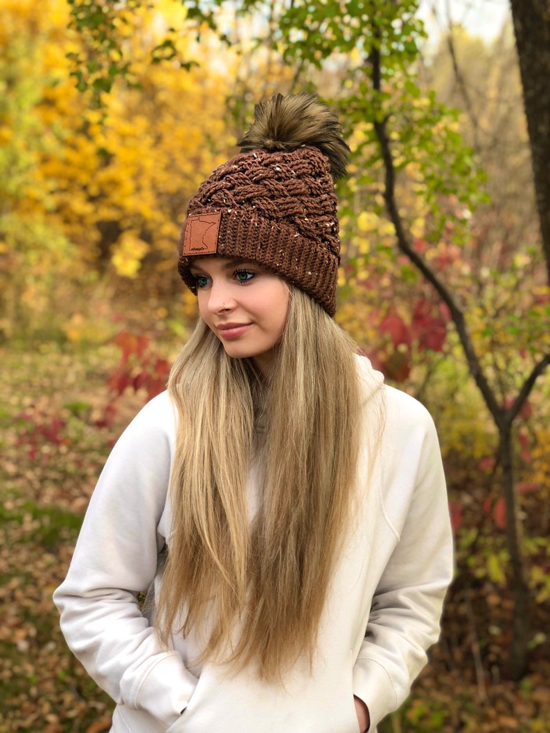 K&ouml;nnte beinhalten: Eine braune Strickm&uuml;tze mit einem Kunstfellbommel und einem braunen Lederpatch mit der Umrandung des Bundesstaates Minnesota. Die M&uuml;tze wird von einer Person mit langen blonden Haaren und einem wei&szlig;en Hoodie getragen.