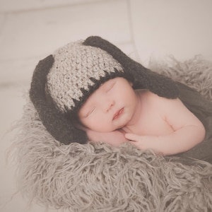 May include: A newborn baby wearing a gray and black bunny hat, sleeping peacefully. The baby is nestled in a soft, gray, fluffy blanket. The hat has long ears, and the baby's skin is smooth and pink.