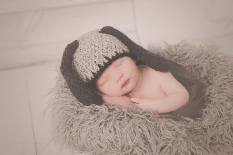 May include: A newborn baby wearing a gray and black bunny hat, sleeping peacefully. The baby is nestled in a soft, gray, fluffy blanket. The hat has long ears, and the baby's skin is smooth and pink.