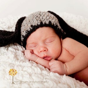 May include: A newborn baby wearing a handmade crochet dog hat with long floppy ears, lying peacefully on a soft, white, textured blanket. The hat is black and gray. The baby is wearing a diaper.