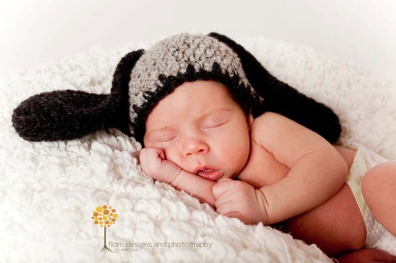 May include: A newborn baby wearing a handmade crochet dog hat with long floppy ears, lying peacefully on a soft, white, textured blanket. The hat is black and gray. The baby is wearing a diaper.