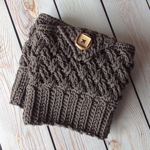 Puede incluir: Polainas de punto de ganchillo marrón con un botón de madera. Las polainas están dobladas y colocadas sobre una superficie de madera.