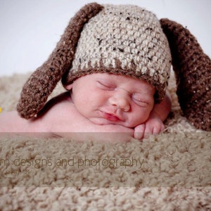 May include: A newborn baby wearing a handmade, brown and tan crochet bunny hat with long ears, resting on a soft, textured blanket. The baby is sleeping peacefully. The image includes the text "flom designs and photography".