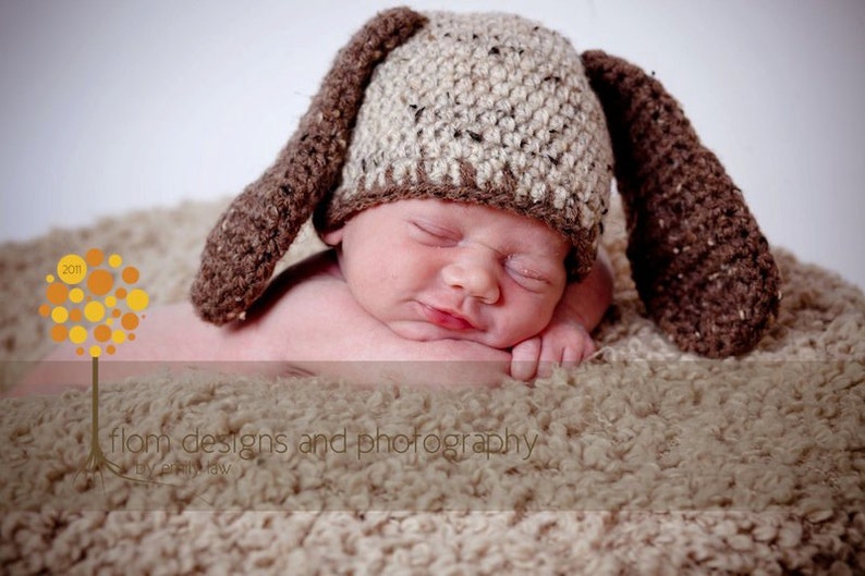 May include: A newborn baby wearing a handmade, brown and tan crochet bunny hat with long ears, resting on a soft, textured blanket. The baby is sleeping peacefully. The image includes the text "flom designs and photography".