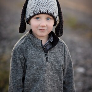 May include: A child wearing a handmade crochet hat with long, floppy black ears. The hat is gray with a dark brown trim. The child is also wearing a gray zip-up sweater.