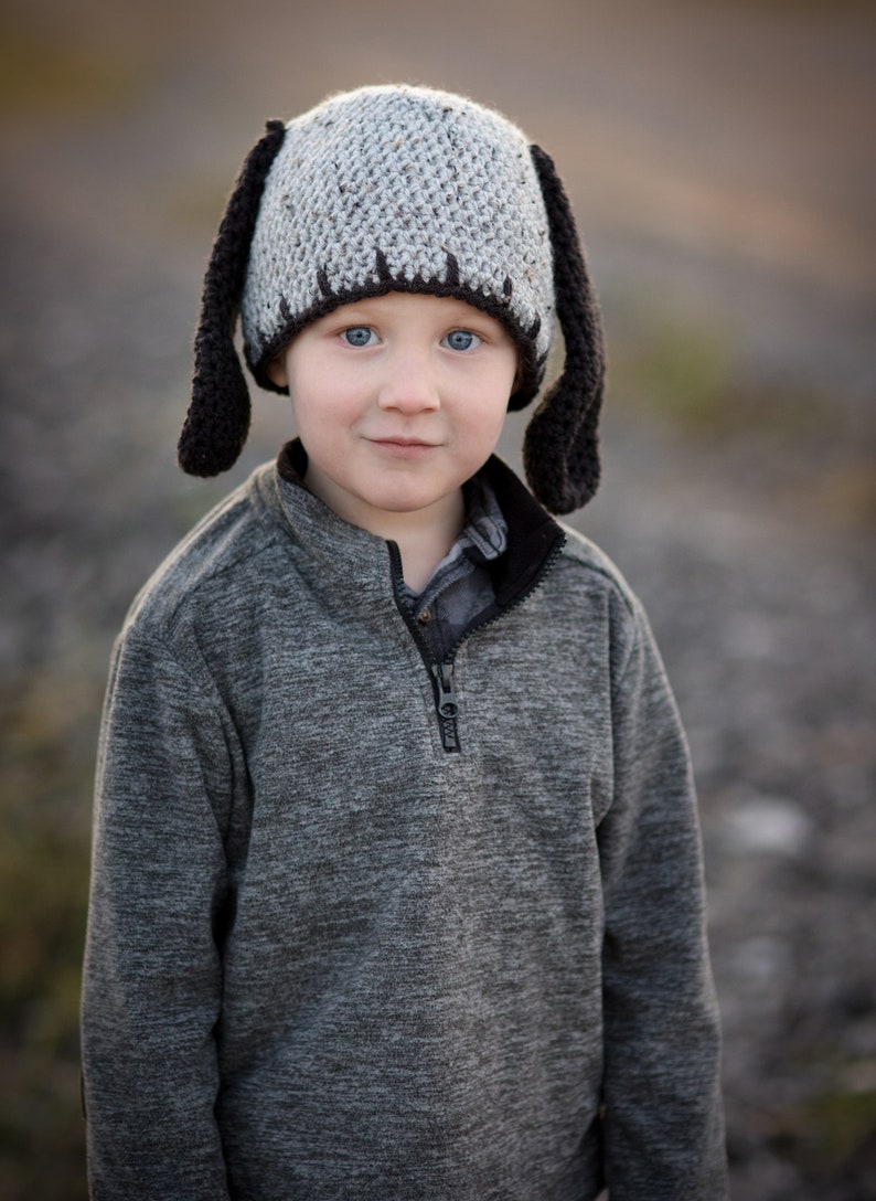 May include: A child wearing a handmade crochet hat with long, floppy black ears. The hat is gray with a dark brown trim. The child is also wearing a gray zip-up sweater.