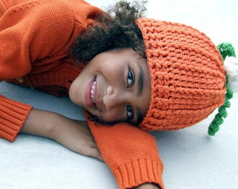 May include: A young person wearing an orange knitted sweater and a crocheted pumpkin hat with a green stem and white flower accents.