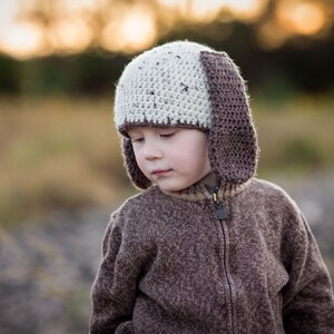 May include: A child wearing a handmade crochet hat with floppy ears and a matching brown zip-up sweater. The hat is two-toned, with a light beige crown and dark brown ear flaps. The sweater is a solid brown color.