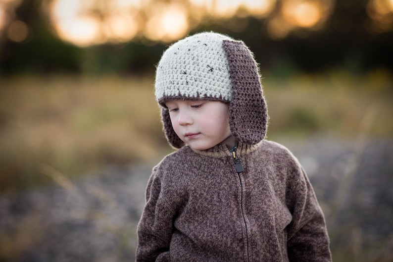 May include: A child wearing a handmade crochet hat with floppy ears and a matching brown zip-up sweater. The hat is two-toned, with a light beige crown and dark brown ear flaps. The sweater is a solid brown color.