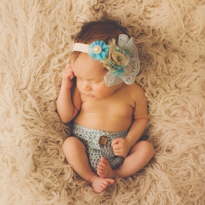May include: A baby girl wearing a light blue crocheted diaper cover with a brown button and a headband with a blue and brown flower.