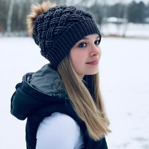 K&ouml;nnte beinhalten: Eine Frau tr&auml;gt eine graue Strickm&uuml;tze mit einem flauschigen Bommel, ein wei&szlig;es Langarmshirt und eine schwarze Weste. Sie steht auf einem schneebedeckten Feld.