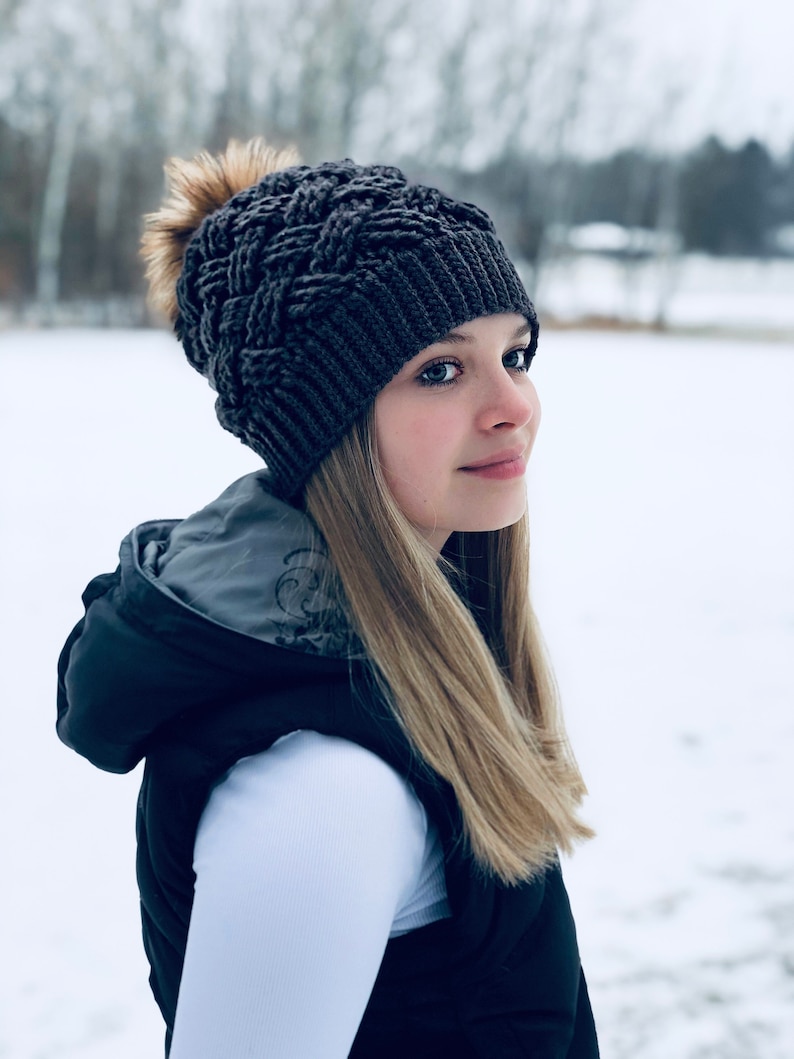 K&ouml;nnte beinhalten: Eine Frau tr&auml;gt eine graue Strickm&uuml;tze mit einem flauschigen Bommel, ein wei&szlig;es Langarmshirt und eine schwarze Weste. Sie steht auf einem schneebedeckten Feld.