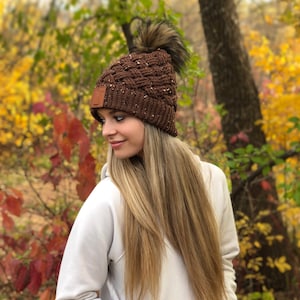 K&ouml;nnte beinhalten: Eine braune Strickm&uuml;tze mit einem Kunstfellbommel. Die M&uuml;tze hat ein geflochtenes Muster und ein kleines braunes Lederst&uuml;ck an der Seite.