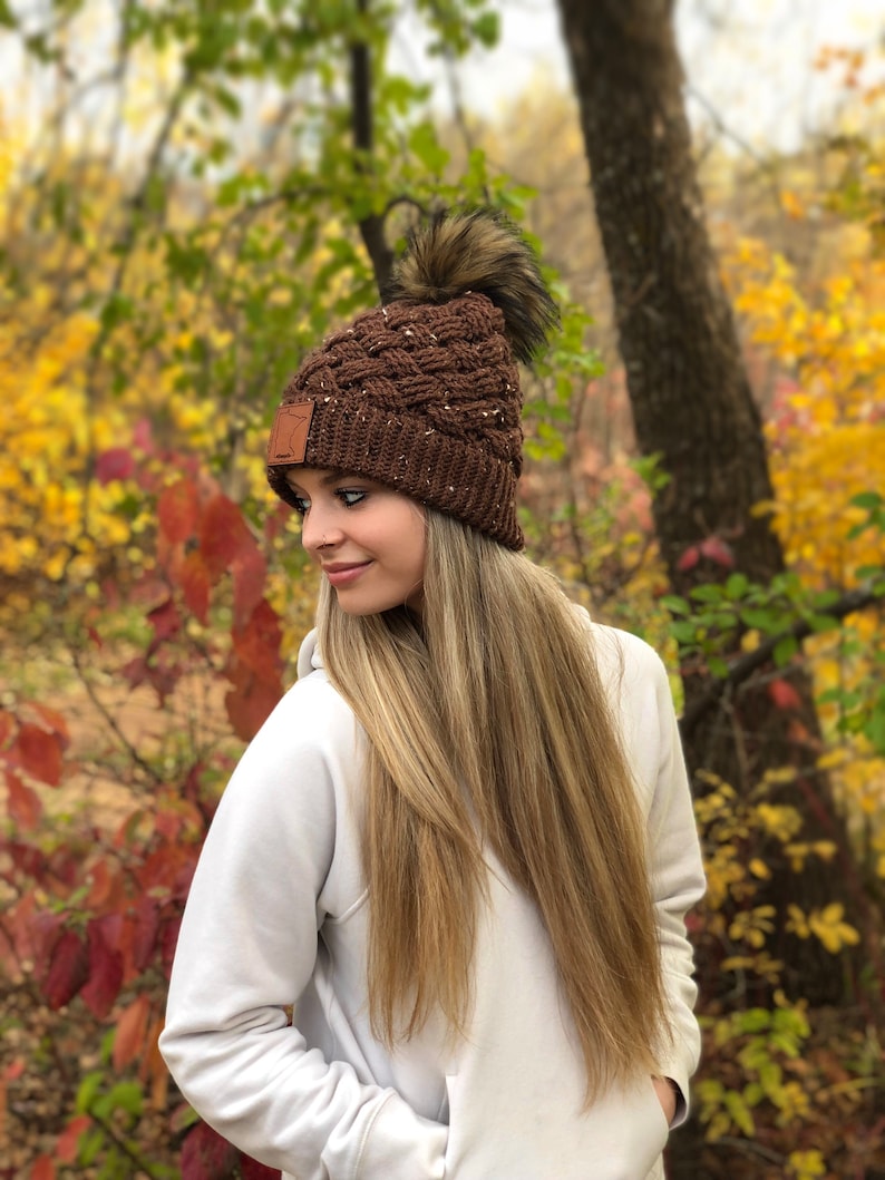 K&ouml;nnte beinhalten: Eine braune Strickm&uuml;tze mit einem Kunstfellbommel. Die M&uuml;tze hat ein geflochtenes Muster und ein kleines braunes Lederst&uuml;ck an der Seite.