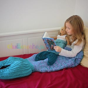 Op de afbeelding: Een gehaakte zeemeerminstaartdeken in blauw- en groentinten. Een meisje en een pop lezen een boek op een rood bed. De deken heeft een vinnenachtig ontwerp aan het einde.