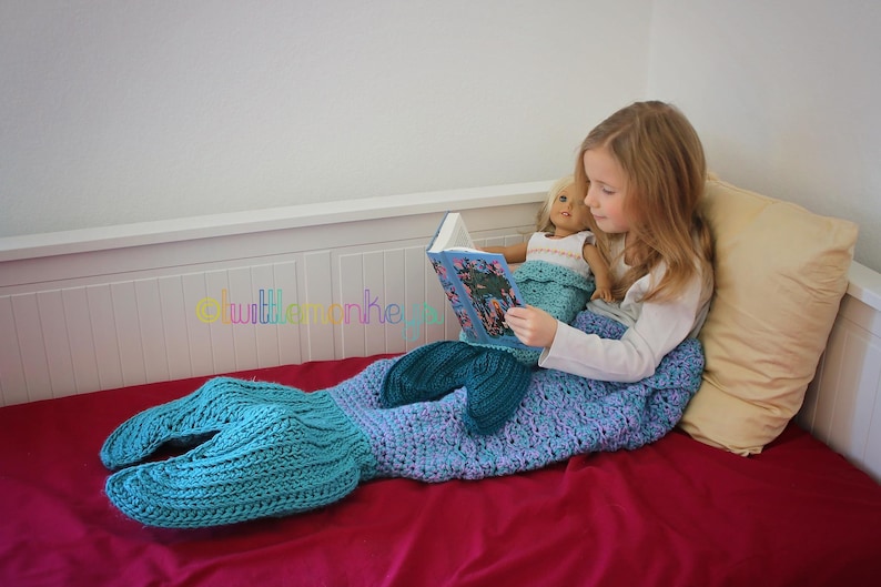 Op de afbeelding: Een gehaakte zeemeerminstaartdeken in blauw- en groentinten. Een meisje en een pop lezen een boek op een rood bed. De deken heeft een vinnenachtig ontwerp aan het einde.